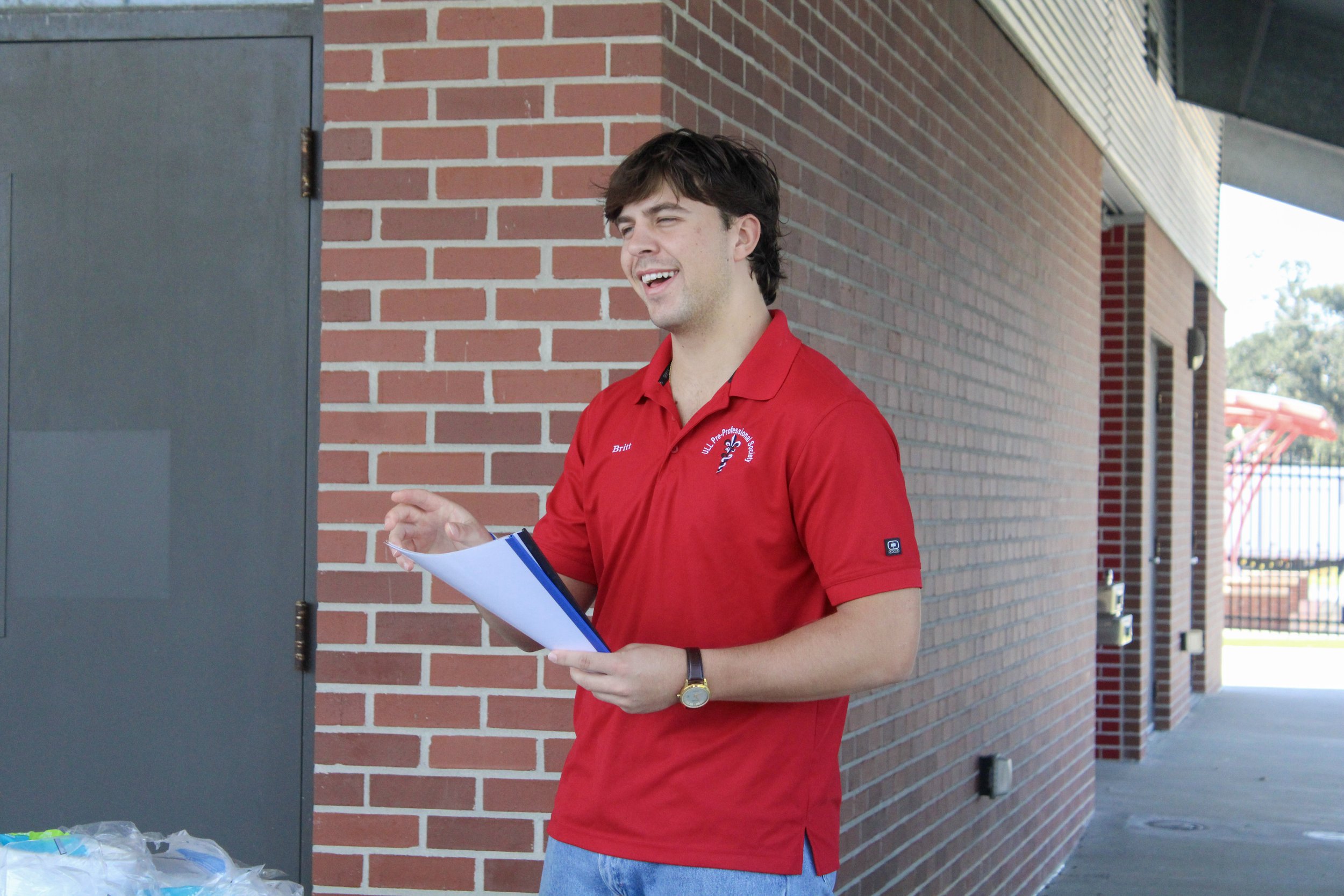 A young man with brown hair smiling while holding papers, wearing a red polo shirt with a logo on the chest, standing next to a brick wall outdoors.