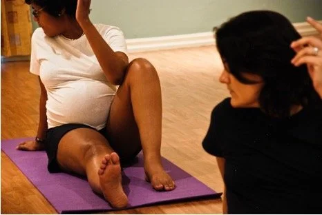 Two women engaging in yoga or stretching exercises in a room with wooden floors, one sitting on a purple yoga mat and the other sitting nearby.
