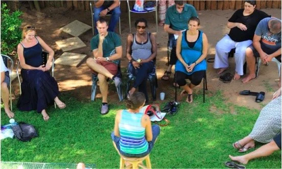 Group of people sitting outdoors, attentively listening to a woman speaking in front of them.