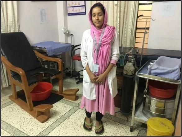 Young woman in a pink dress and headscarf standing in a room, possibly a medical facility, with hospital equipment and furniture around.