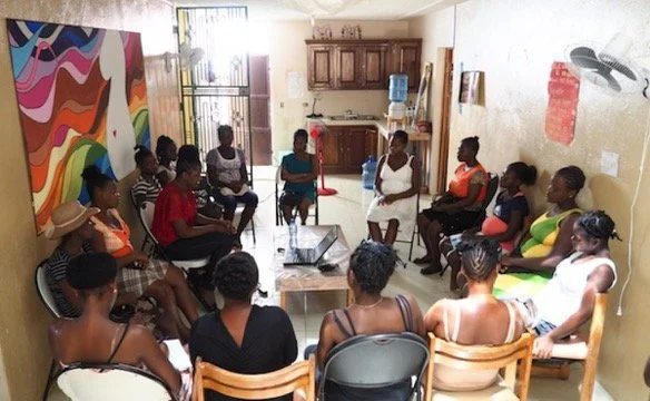 Women sitting in a circle during a group meeting in a living room with a colorful mural on the wall.