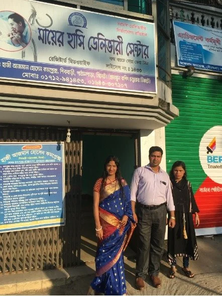 Three people standing outside a building with a signboard above and a blue information board on the wall. Two women and one man, dressed in semi-formal attire, are posing for the photo.