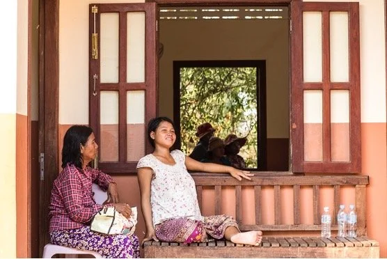 Two women sitting on a wooden ledge outside a building with open windows, with one woman smiling and the other looking at her; there are three water bottles on the ledge and trees visible through the window.