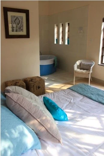 Bedroom with a bed covered in white and teal bedding, a blue pillow, a wicker chair, some pillows on the bed, a framed artwork on the wall, and a hot tub in the corner near three small windows.