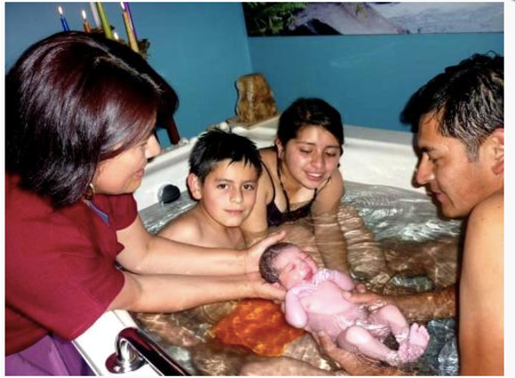 Family of four, including two children and a newborn, in a hot tub or small pool, with a woman in scrubs assisting, indoors.