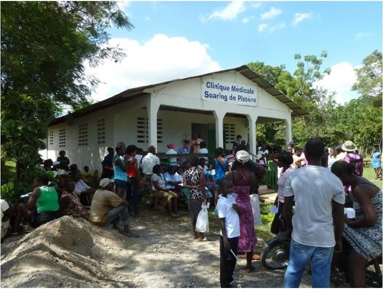 Many people gathered outside a small medical clinic with a sign reading 'Clinique Medicale Soaring de Pister' in a rural area, with some sitting on benches and others standing near the entrance.