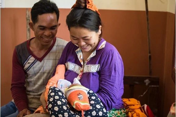 A woman wearing purple and an orange headscarf holding a baby with a hat, while a man stands next to her, all smiling.