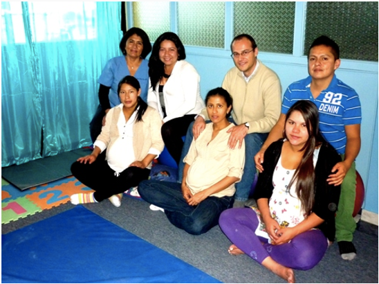 Group of seven people, including four women and three men, sitting and standing in a room with blue curtains and colorful foam mats on the floor.