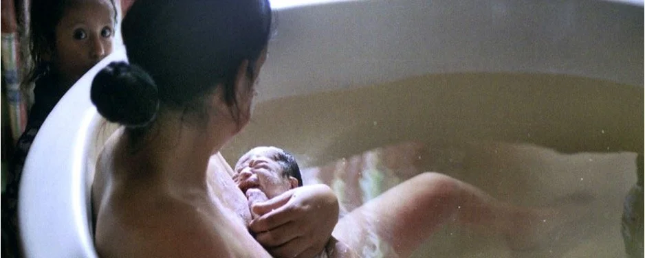 Woman giving birth in a birthing pool with two children observing nearby