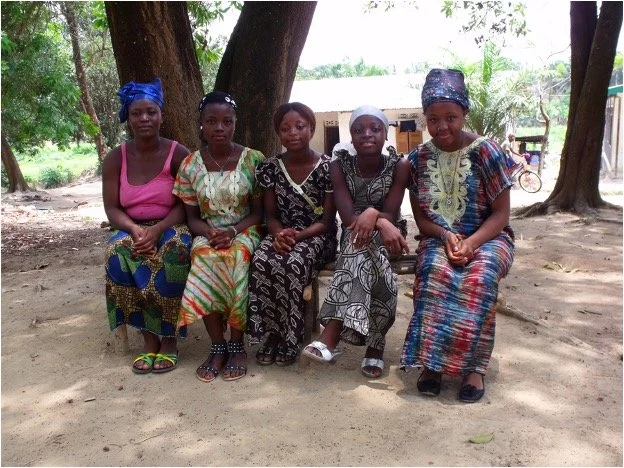 Five women sitting on a bench outdoors under trees in a rural area.