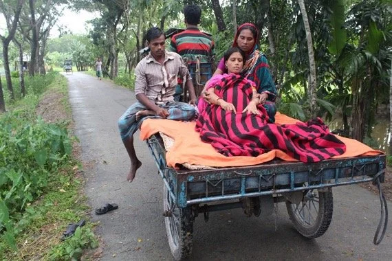A woman sitting on a cart with a blanket, accompanied by two men and a woman nearby, on a rural road surrounded by trees.