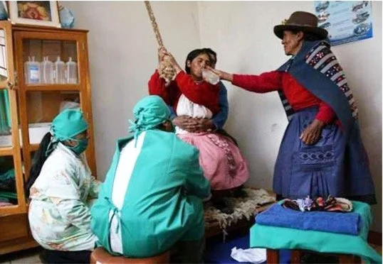 A woman receiving a traditional beverage from another woman in a room with medical supplies, with two other women sitting nearby.