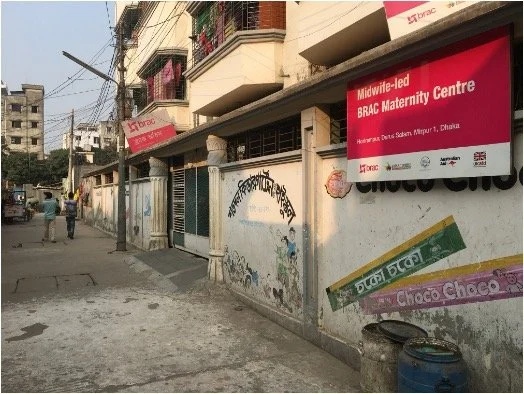 Street view in an urban area with buildings, electrical wires, and a few pedestrians, featuring a sign for a Midwifery-led BRAC Maternity Centre.