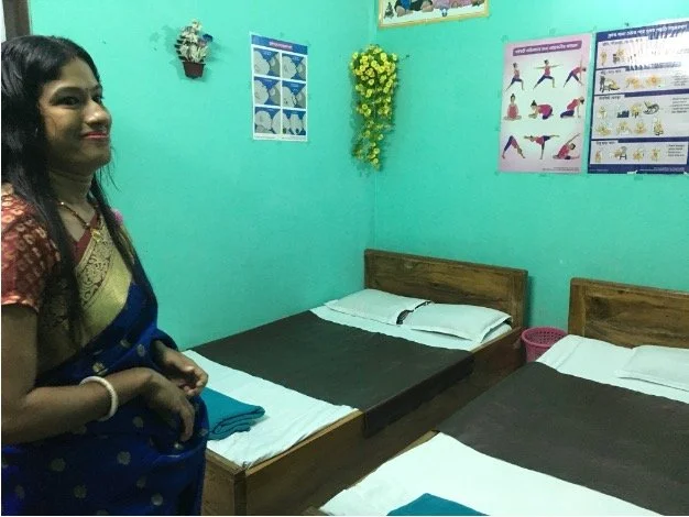 A woman in traditional attire standing in a room with two beds, turquoise walls, and dental health posters on the wall.
