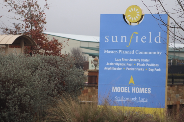 Blue sign for Sunfield community with amenities and model homes, surrounded by trees and bushes