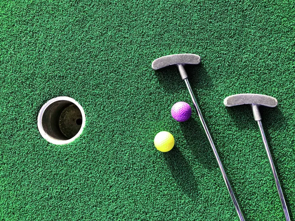 Two mini golf clubs and two golf balls, one purple and one yellow, on a green mini golf course with a hole visible.