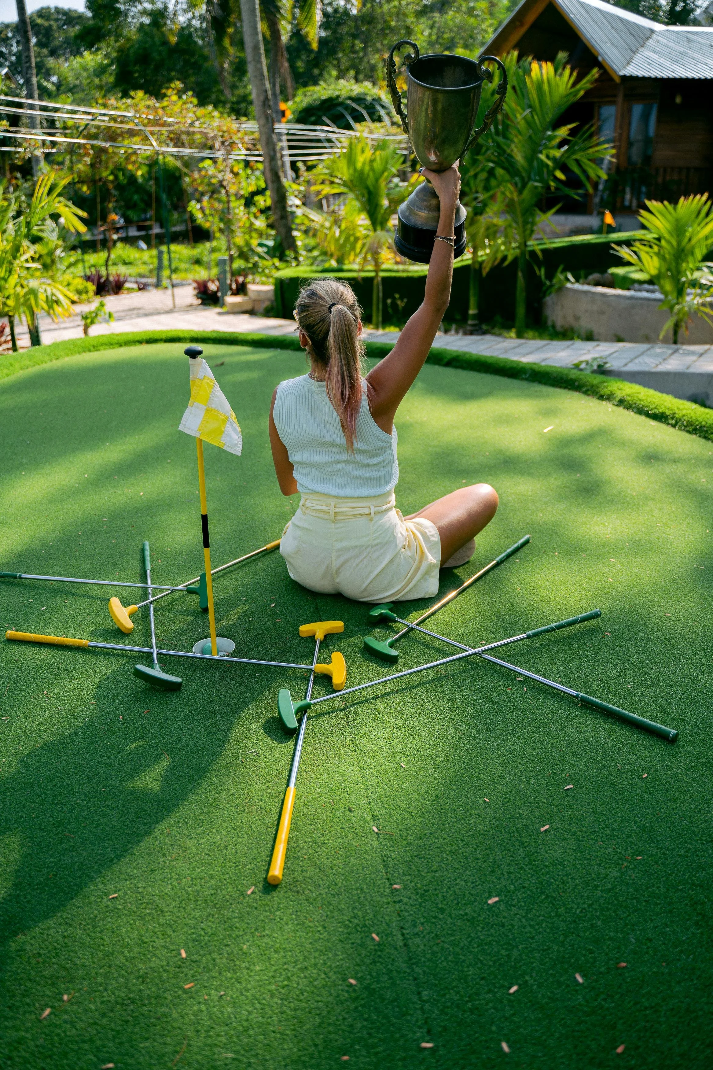 A young woman sitting on a mini golf course green, holding a trophy above her head, surrounded by scattered golf clubs. She is wearing a white sleeveless top and light-colored shorts, with a flagstick in front. There are trees, plants, and a small house in the background.