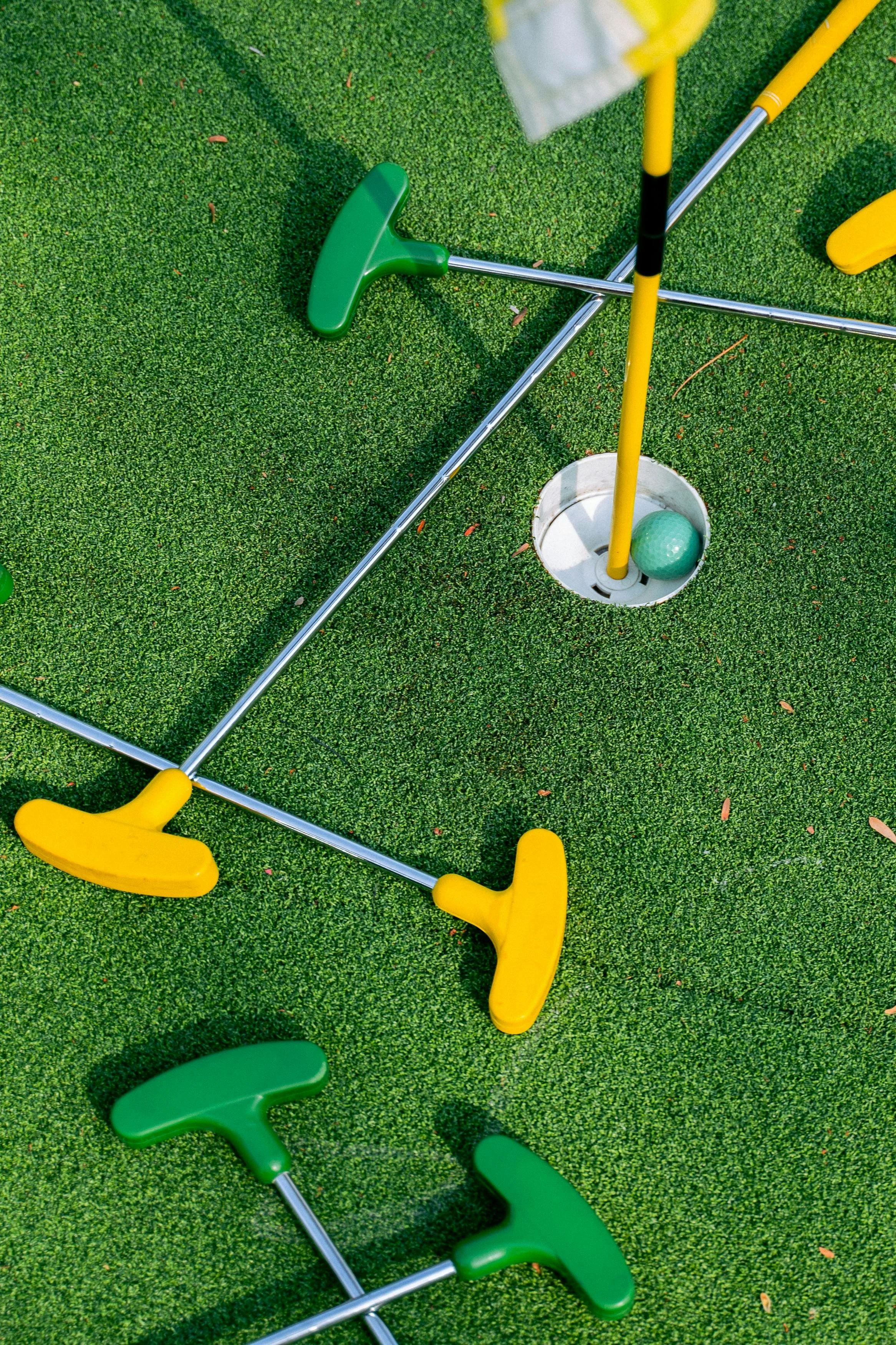 Two miniature golf putters, one green and one yellow, lying on a green artificial turf putting green with a golf ball in the hole near the yellow putter.