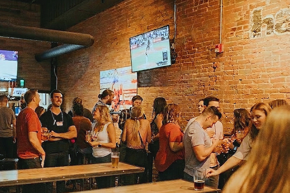 Crowd of people socializing in a bar with exposed brick walls and multiple television screens showing sports.