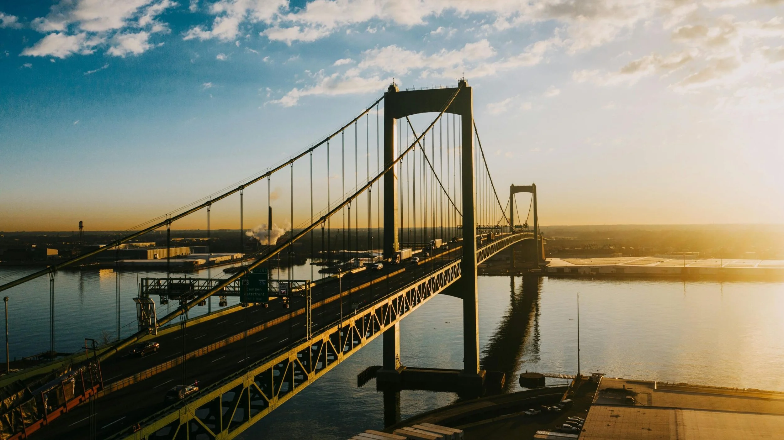 A large suspension bridge spanning over a body of water during sunset, with cars traveling across and a city skyline in the distance.