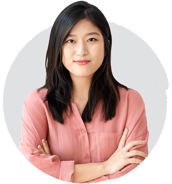 A young woman with long dark hair wearing a pink button-up shirt, crossing her arms and looking at the camera with a neutral expression.