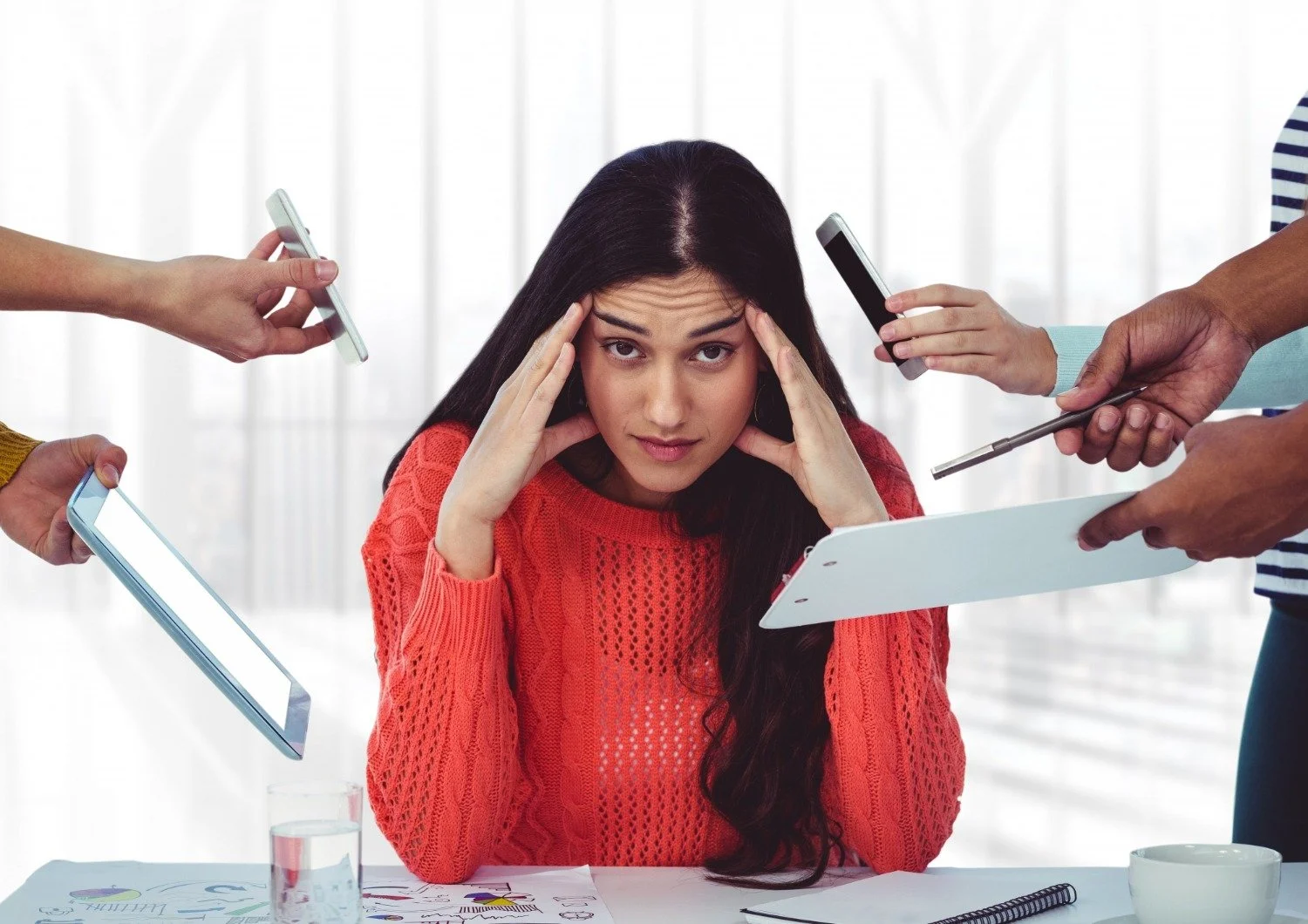 A woman in a red sweater appears stressed as multiple hands with mobile devices and a tablet surround her in an office setting.