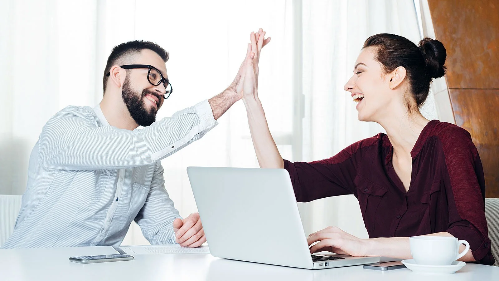 Two colleagues give each other a high five in an office setting, smiling and sitting at a table with a laptop and coffee mug.