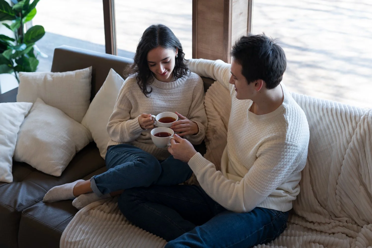 A young woman and man sitting on a beige couch with white pillows, sharing a moment over two cups of tea in a cozy, well-lit room next to a large window with a view of water outside.