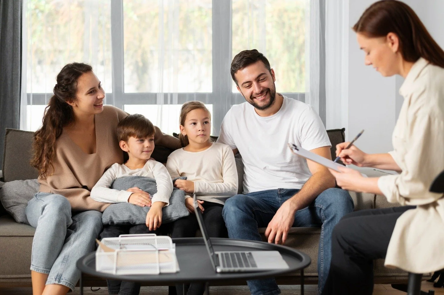 A family of five sitting on a sofa during a meeting with a therapist, with a laptop and documents on a table in front of them. The parents and two children are paying attention to the therapist.