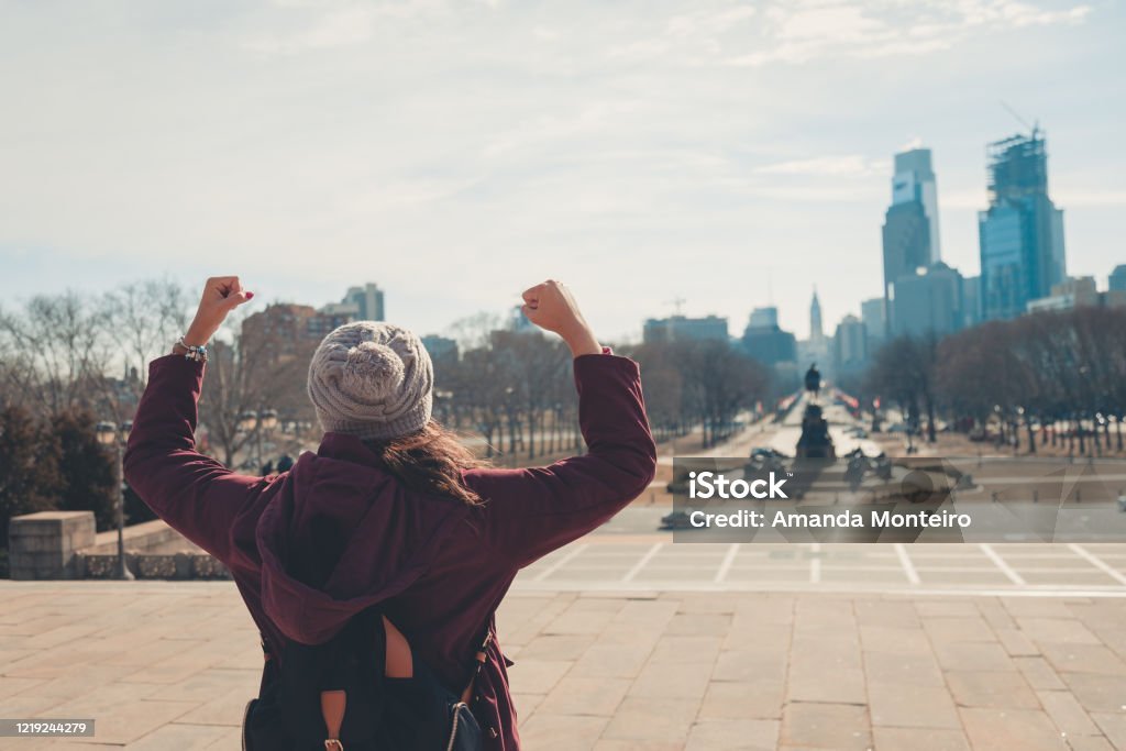 Person wearing a knit beanie and burgundy jacket stands with their back to the camera, flexing their arms in front of a city skyline on a sunny day.
