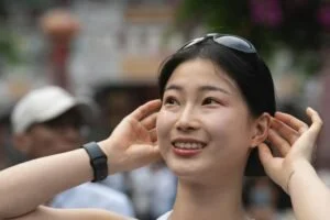 A smiling Asian woman outdoors, with sunglasses on her head, touching her ears, in a crowd.