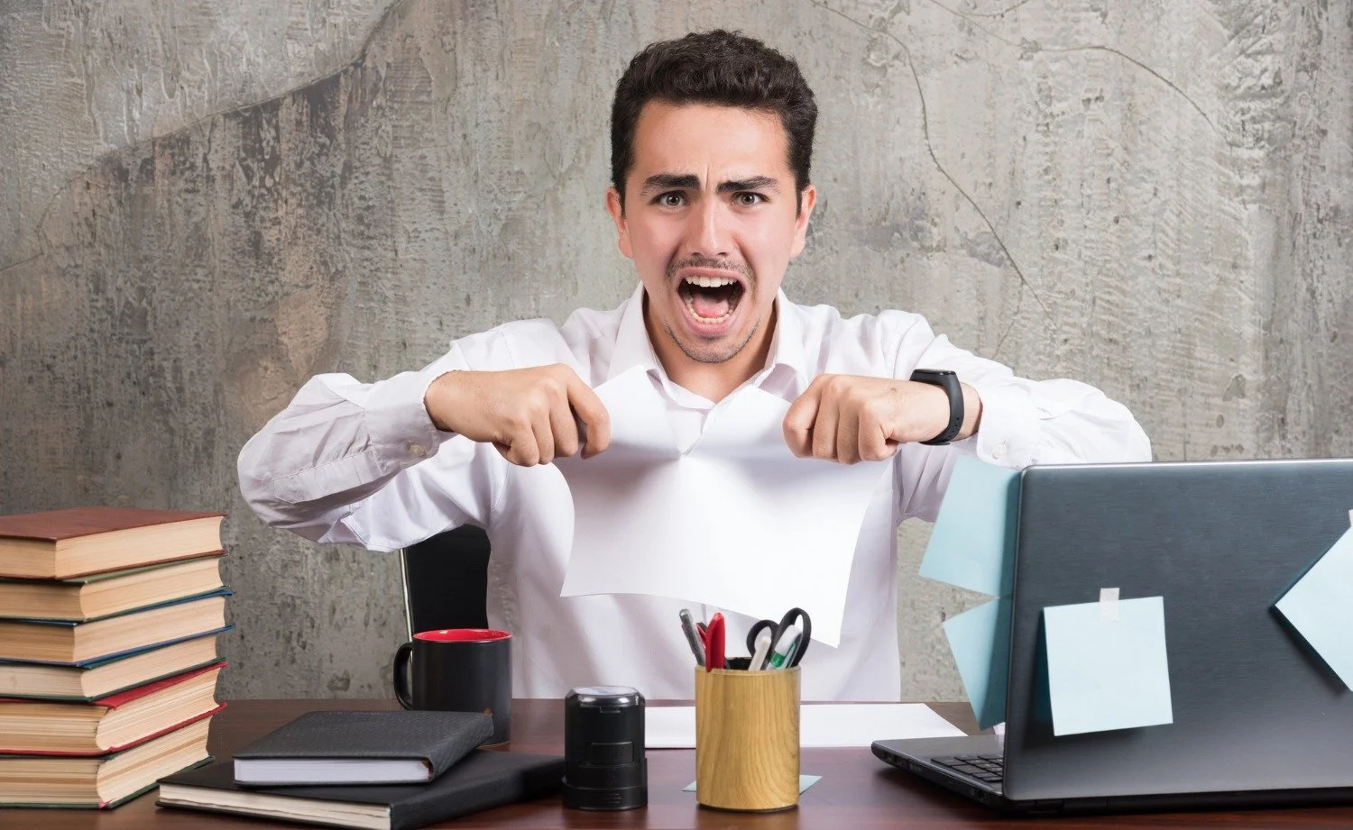 Man in white shirt tearing a sheet of paper in an office setting with books, office supplies, and a laptop on the desk.