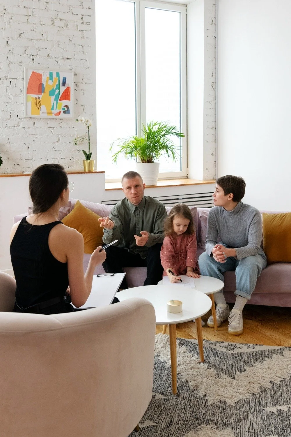 A family of four sitting on a pink sofa in a living room having a discussion with a woman taking notes, two young children, a girl drawing on a small table. The room has a large window, white brick wall, painting, green plant, and cozy decor.