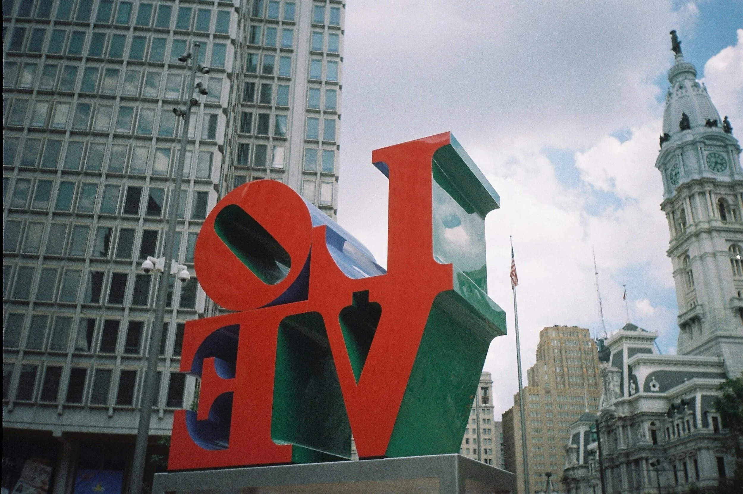 Colorful large sculpture spelling out 'LOVE' with city buildings and a clock tower in the background.
