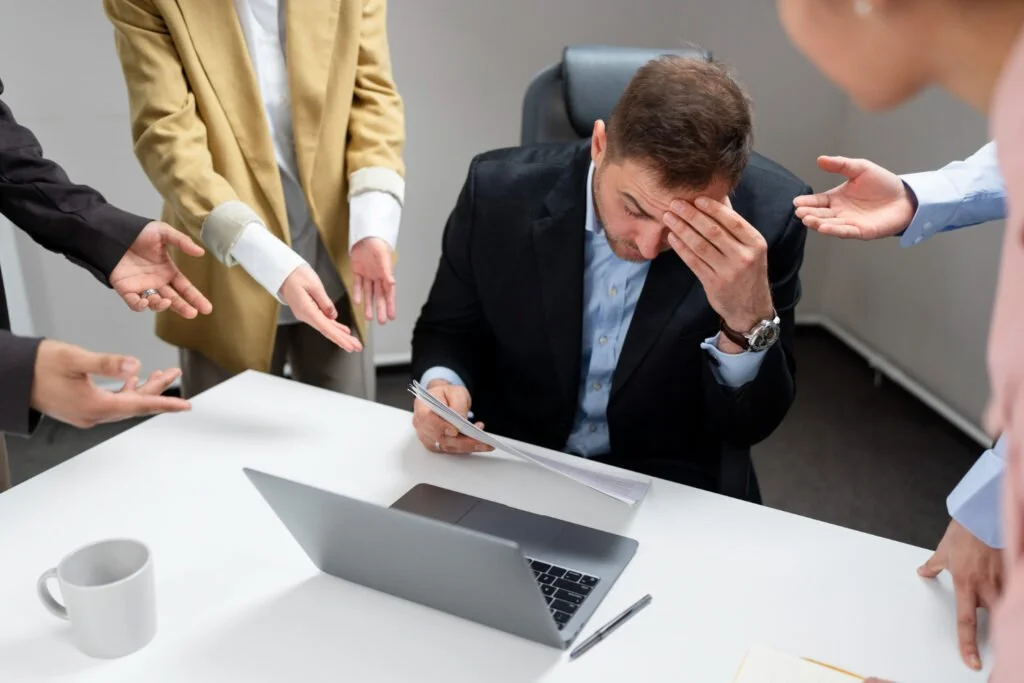 A man in a suit sitting at a desk with a laptop, touching his forehead in distress, surrounded by people gesturing towards him in frustration.