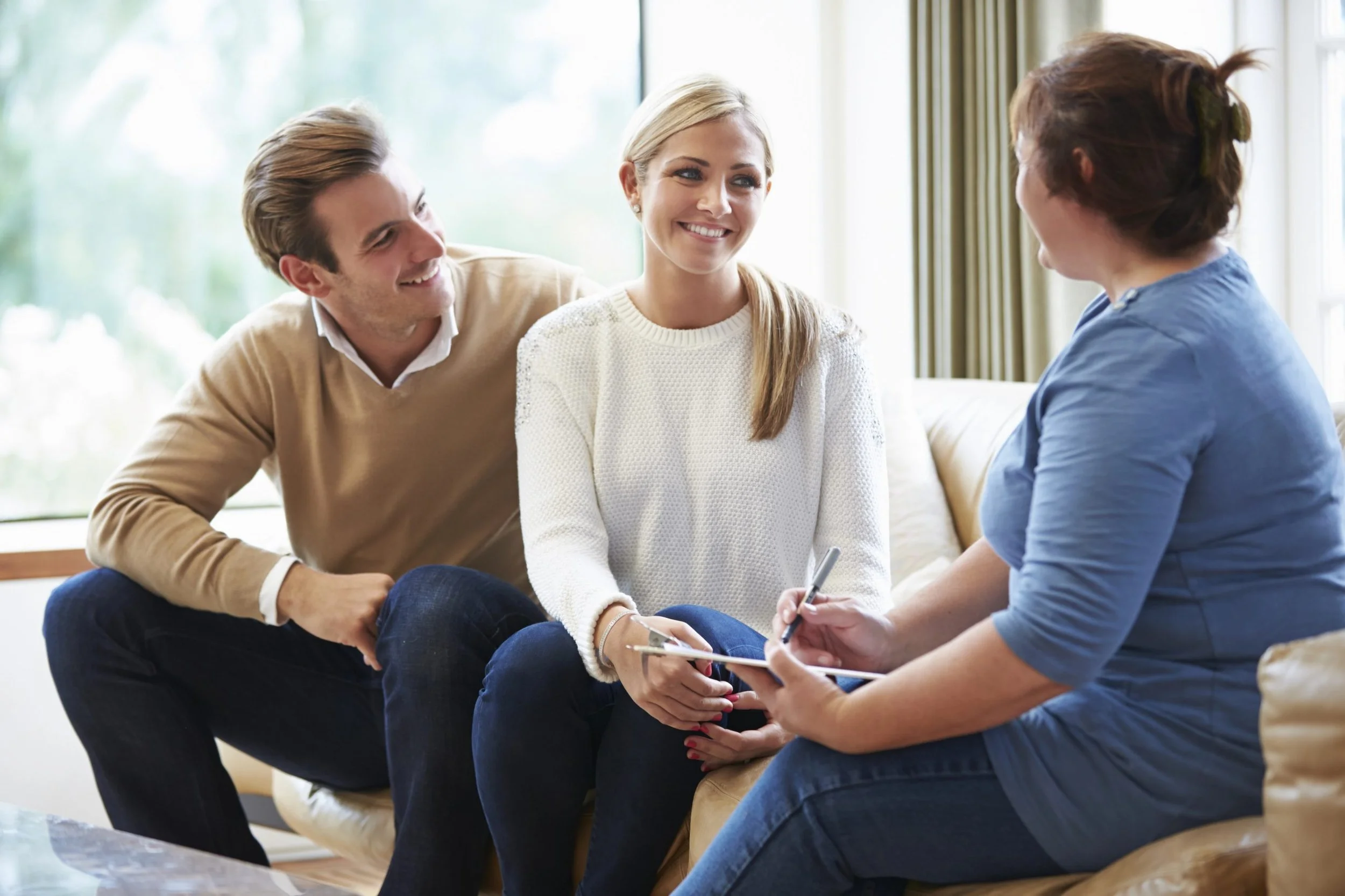 Four people sitting on a sofa, engaged in conversation, with a large window in the background and natural light illuminating the room.