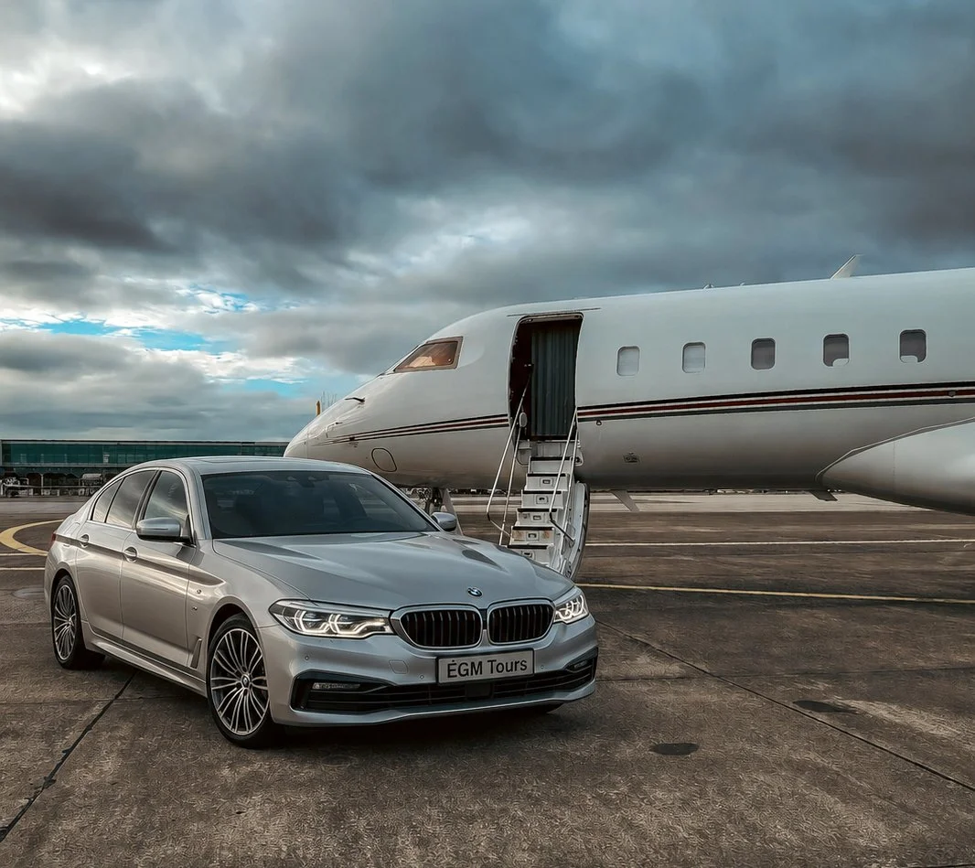 A silver BMW car parked in front of a private jet on an airport tarmac under dark, cloudy skies.