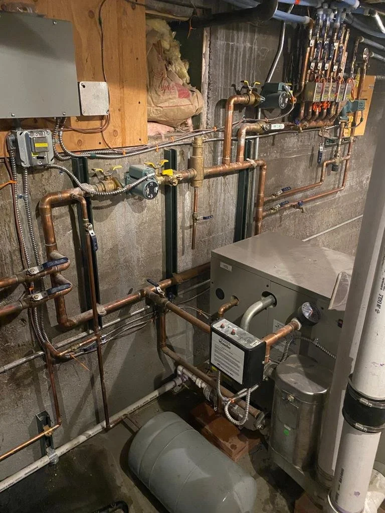 A basement utility room with copper piping, a water heater, a pressure tank, and various valves and gauges.
