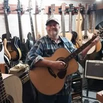 MaCustom Built Guitar Luthier McKiddy Guitarsn in a plaid shirt and baseball cap playing an acoustic guitar in a music store with numerous guitars hanging on the wall behind him.