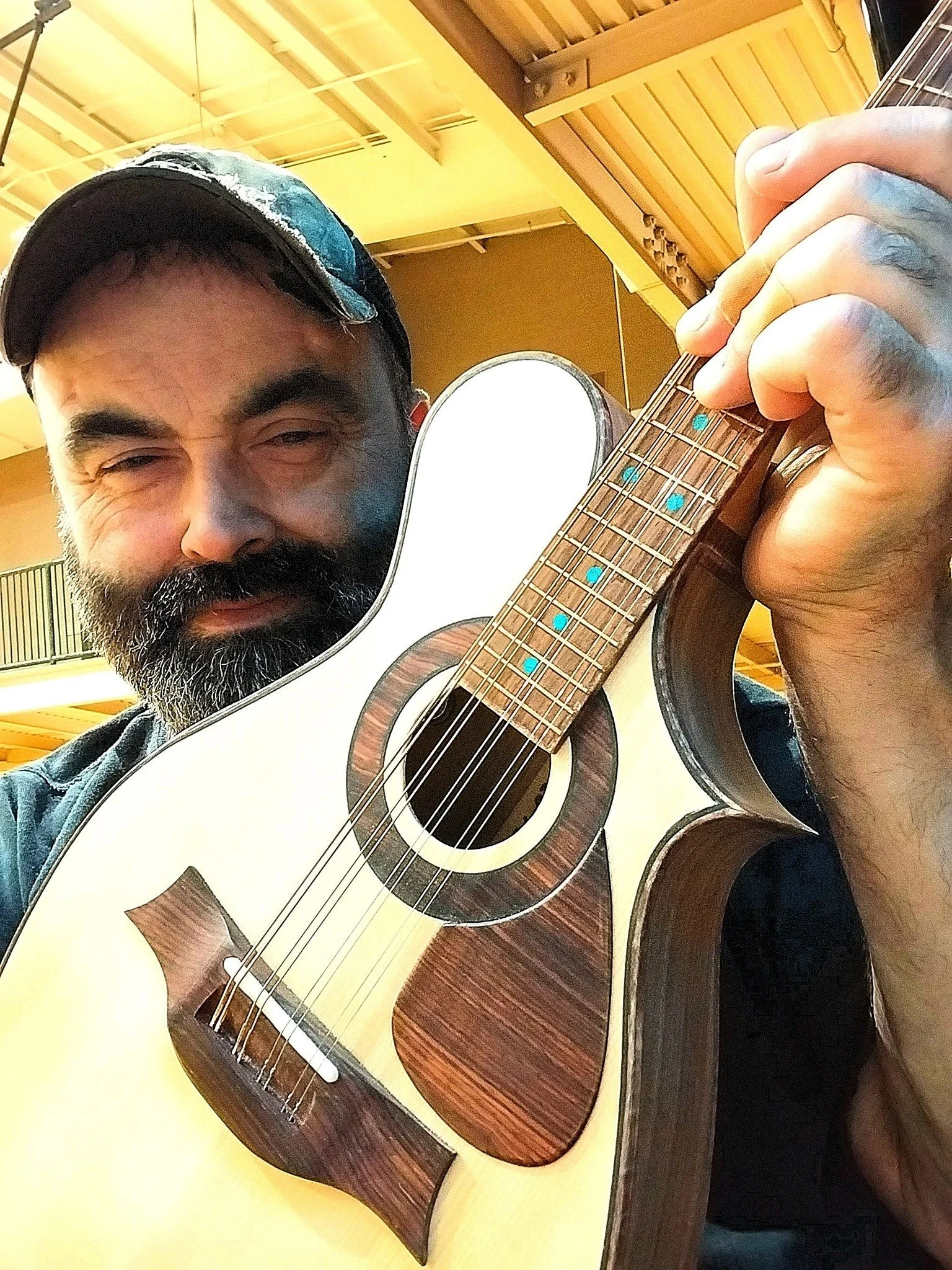 A man with a beard and mustache, wearing a camouflage baseball cap, is holding and playing an acoustic guitar with a natural wood finish in a room with yellow walls and a wooden ceiling.
