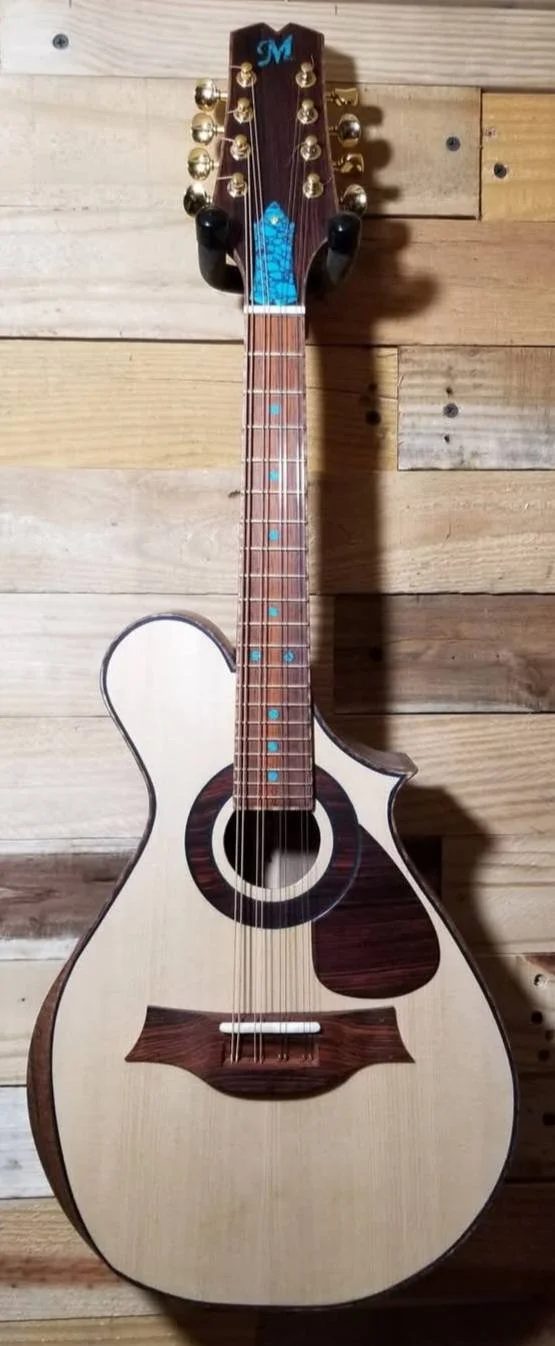 A mandolin with a unique design resembling an acoustic guitar with an extra cutout on the upper left side of the body, placed against a wooden wall.