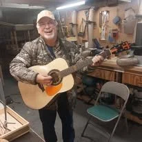 Custom Built Guitar Luthier McKiddy GuitarsMan smiling and playing an acoustic guitar in a workshop or garage.