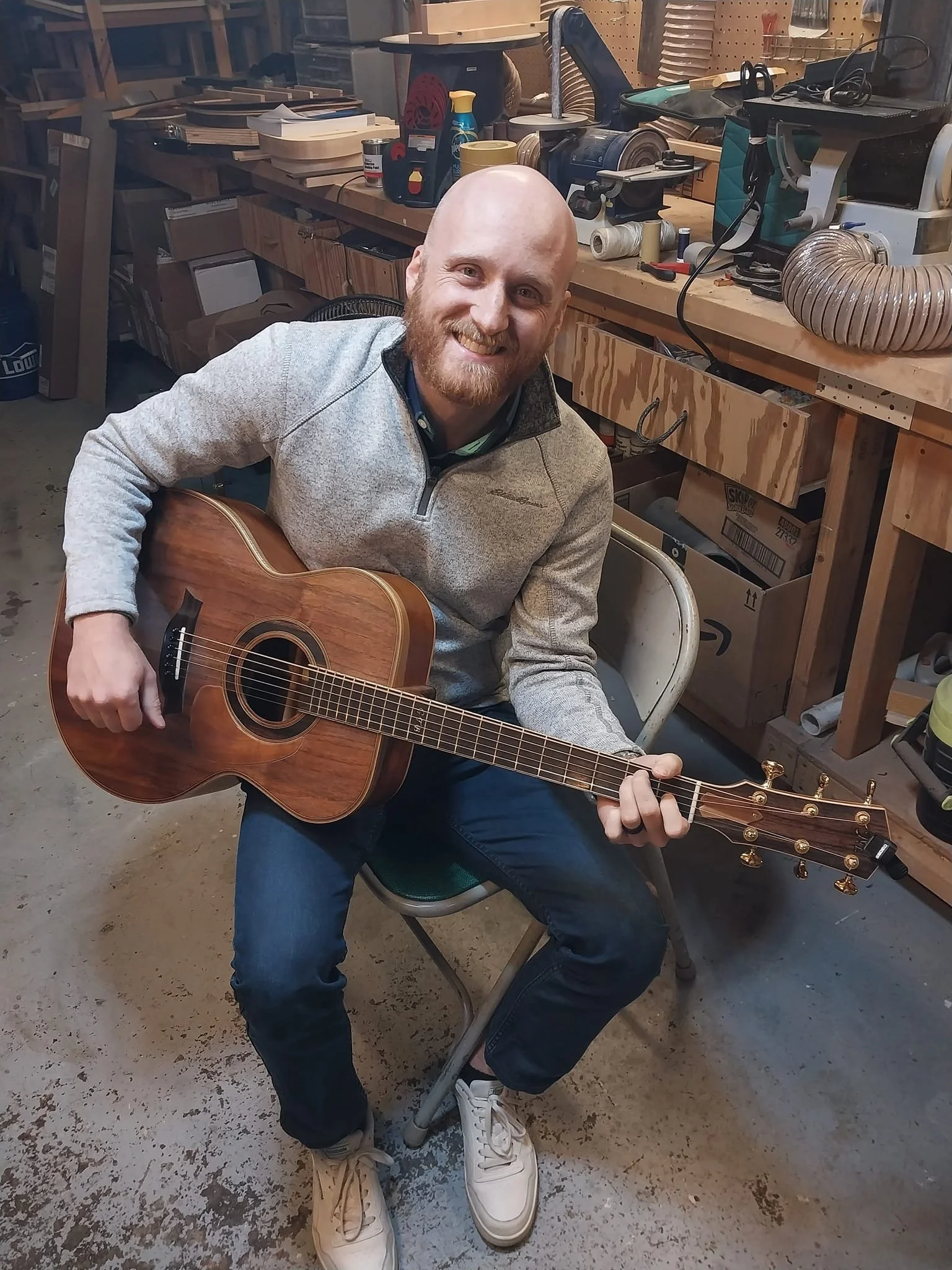 A mCustom Built Guitar Luthier McKiddy Guitarsan with a beard sitting on a chair in a woodworking shop, playing an acoustic guitar. The workshop has a workbench with various tools and equipment in the background.