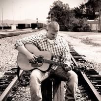 A pCustom Built Guitar Luthier McKiddy Guitarserson sitting on train tracks playing an acoustic guitar.