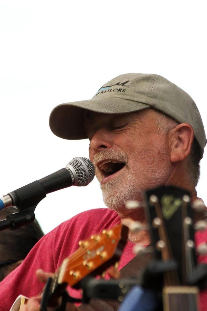 A man singing into a microphone while playing guitar, wearing a tan cap and a red shirt.