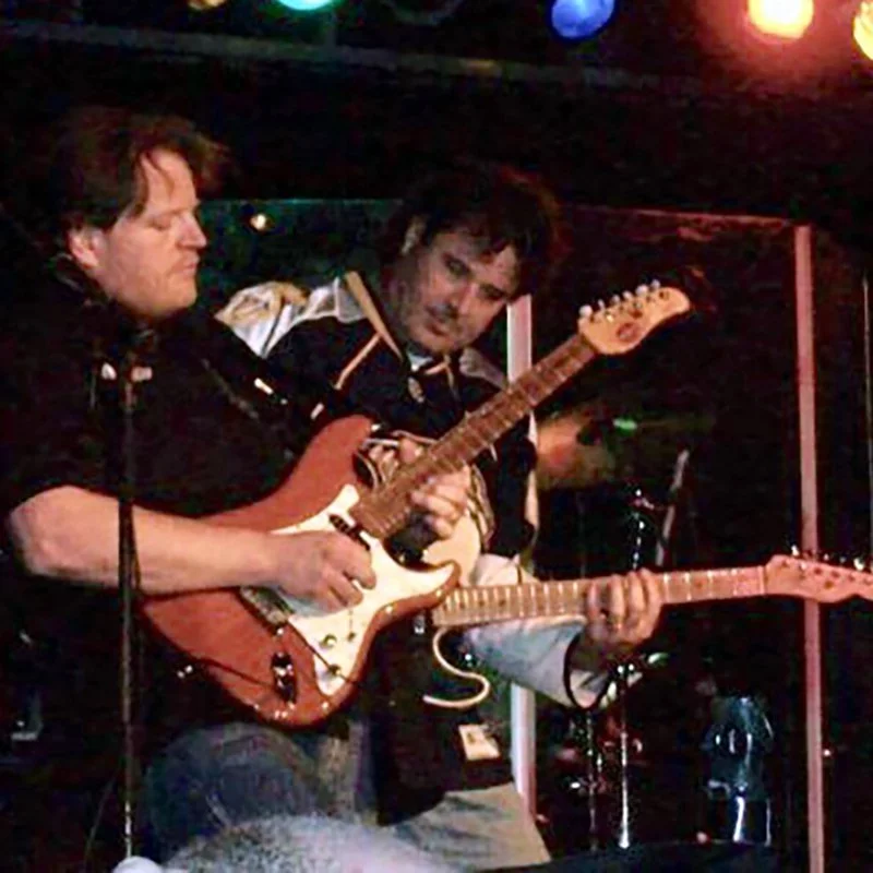 Two men playing electric guitars on a dimly lit stage with colorful lights overhead, focused on their instruments.