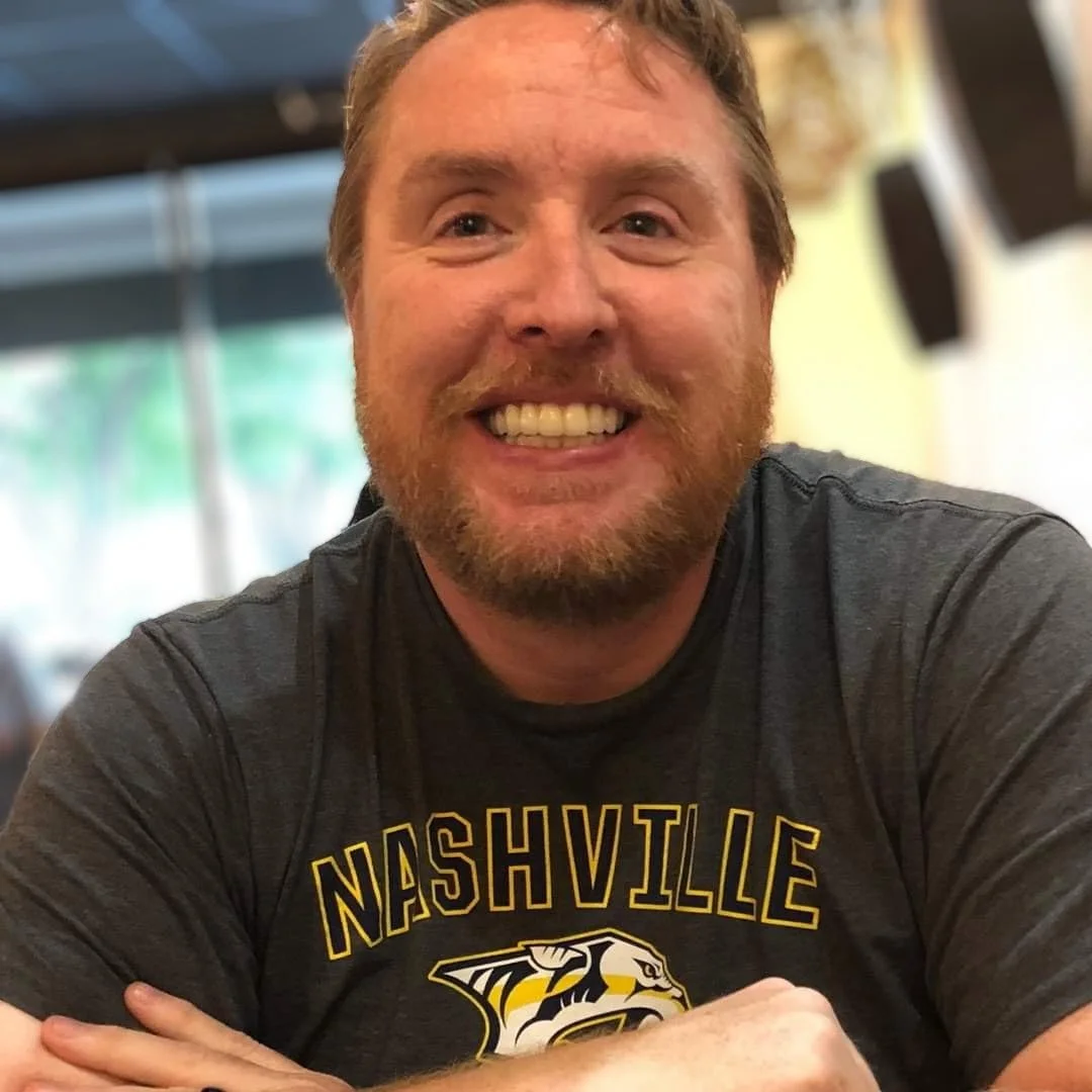 A smiling man with a beard and mustache, wearing a black Nashville sports t-shirt, is sitting indoors with a blurred background.