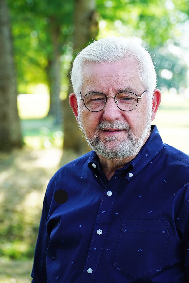 An elderly man with white hair and a beard wearing glasses and a navy blue button-up shirt outdoors in a park with trees and sunlight in the background.