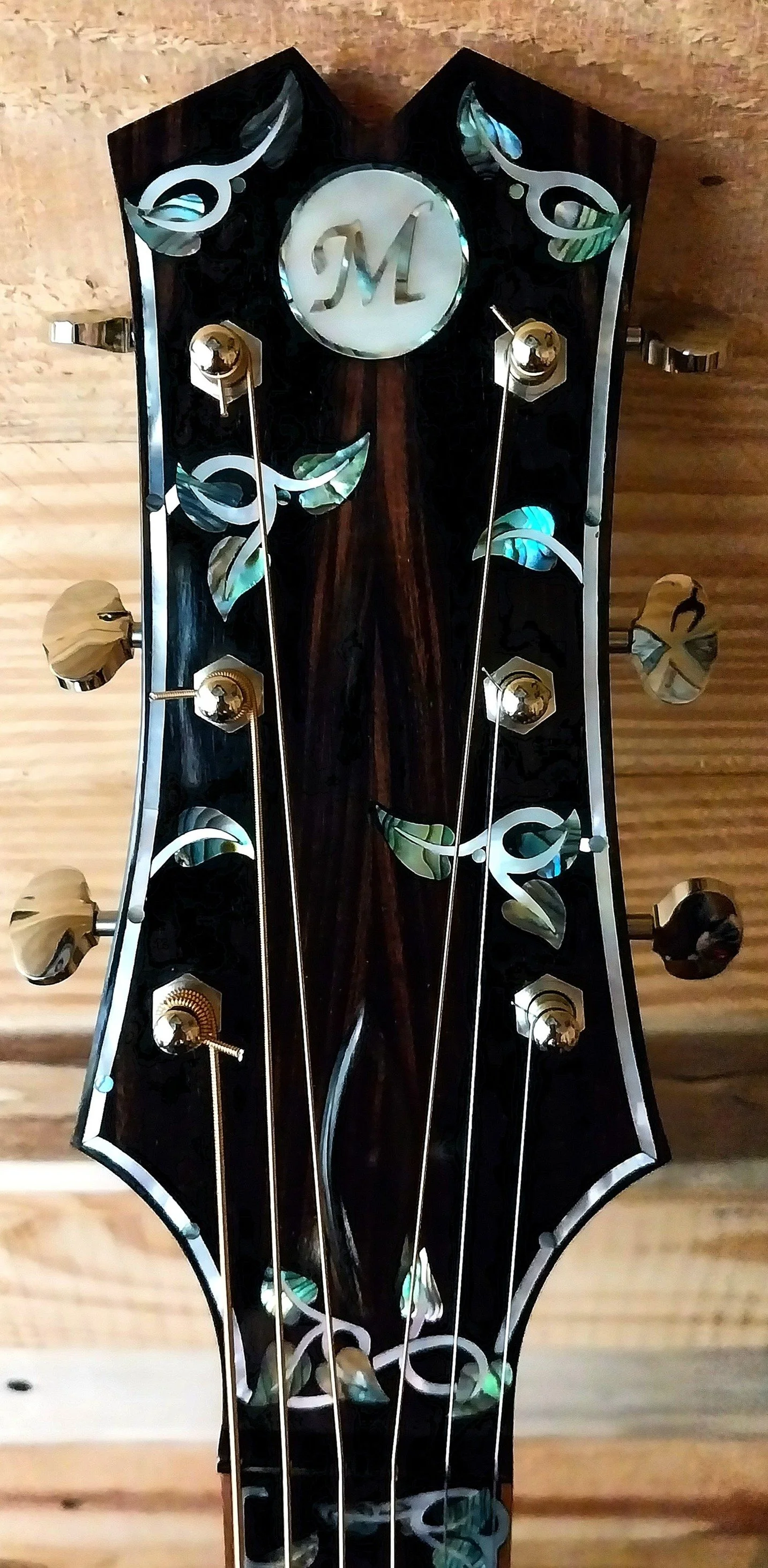 Close-up of a guitar headstock with a dark wood finish, decorative inlays resembling leaves, and a central medallion with the letter 'M' in cursive, set against a wooden background.