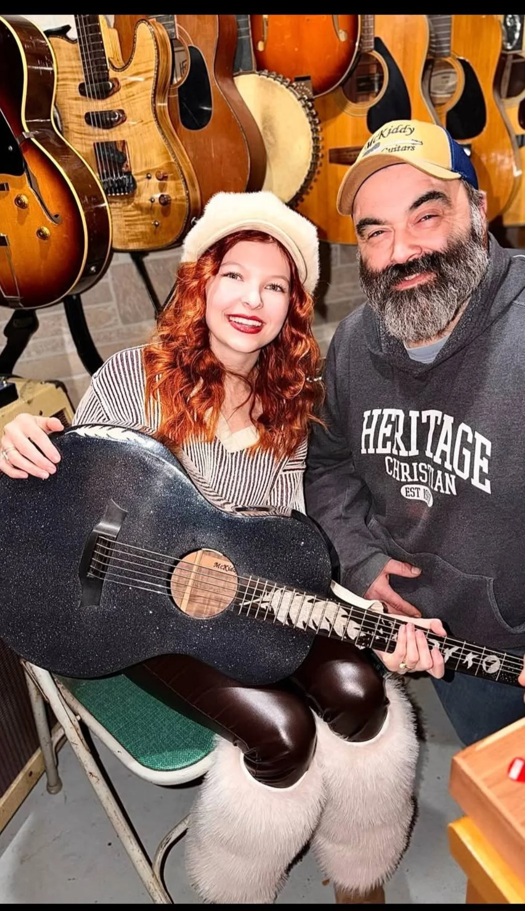 A girl with red hair wearing a white beret holding a black glitter guitar, sitting on a white furry stool, next to a man wearing a gray hoodie and a beige cap, surrounded by multiple guitars on a wall in the background.